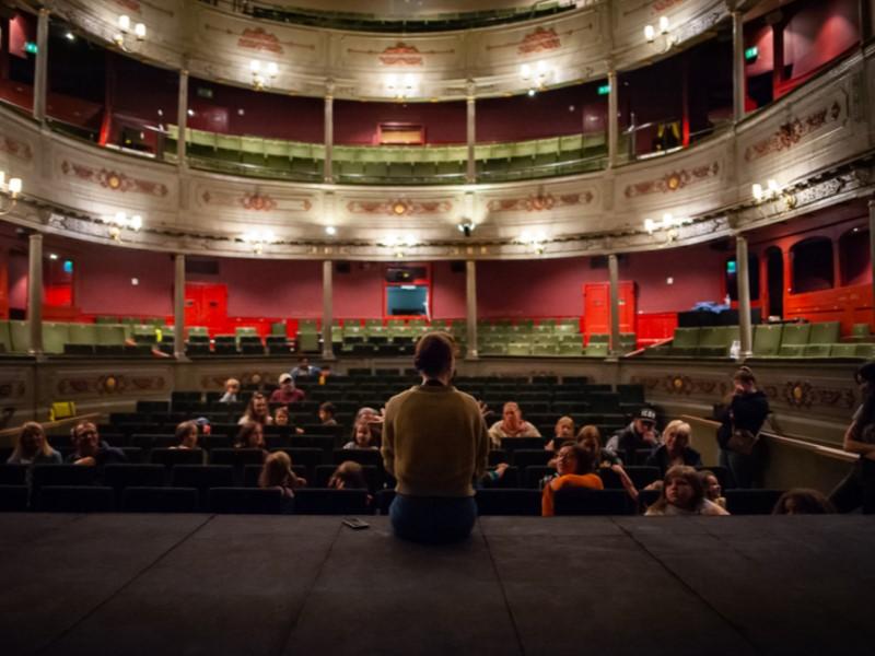 Someone sat on the stage at The Old Vic in Bristol, with their back to camera in front of a crowd of people sat in the stalls.