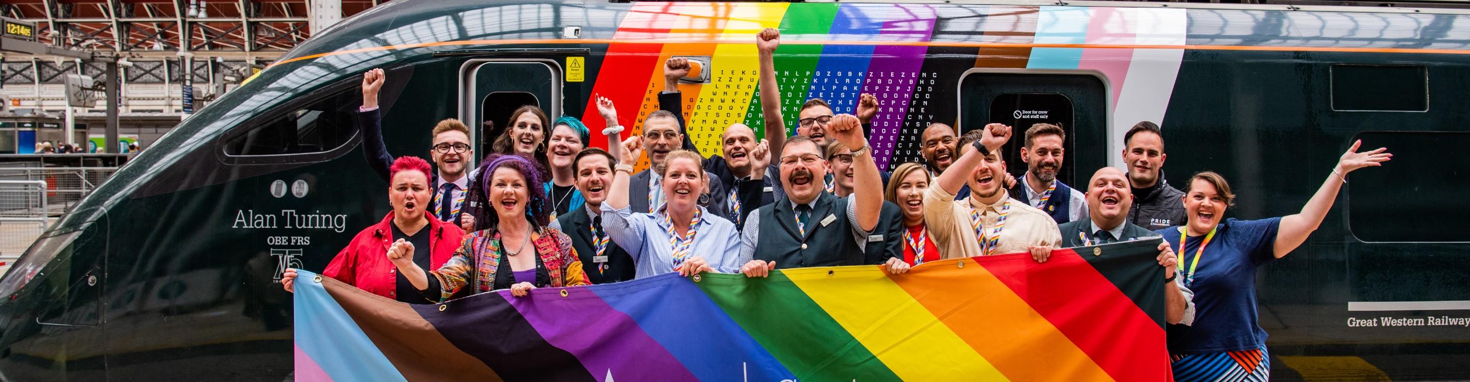 Photo of a cheering crowd on the platform holding a GWR pride rainbow banner in front of the GWR train named for Alan Turing