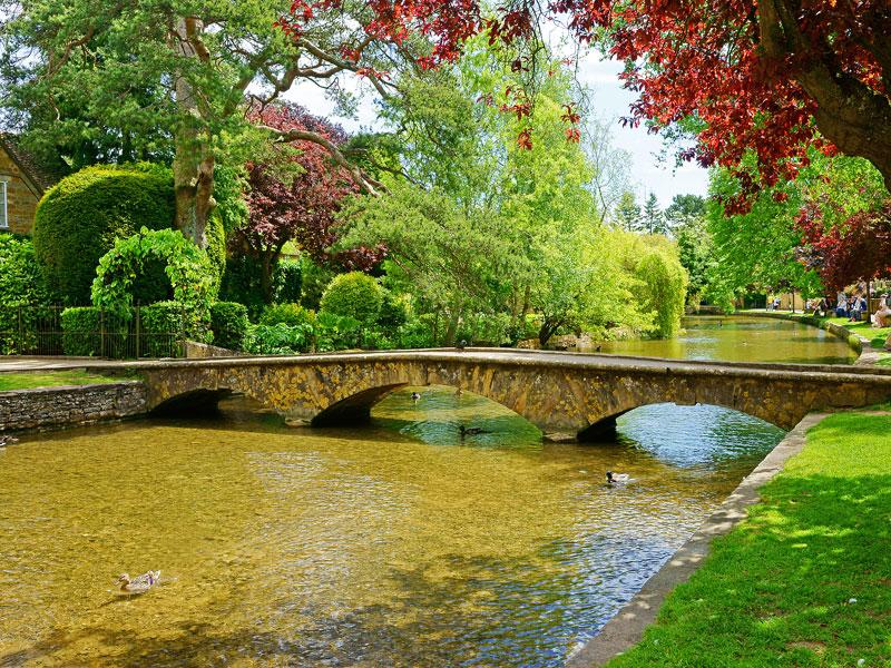The River Windrush flowing through Bourton-on-the-Water, with a low bridge and trees overhanging the water, UK