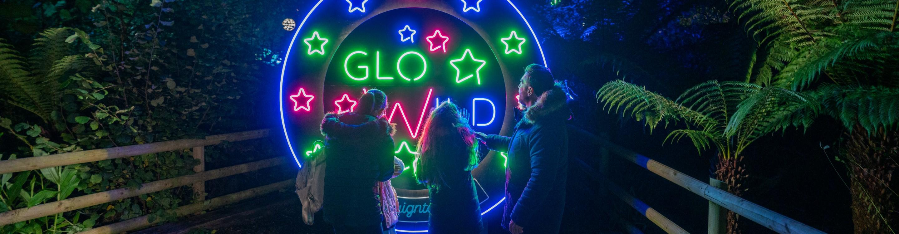 A family at Paignton Zoo at night, looking at the GloWild sign, illuminated in neon blue, green and pink