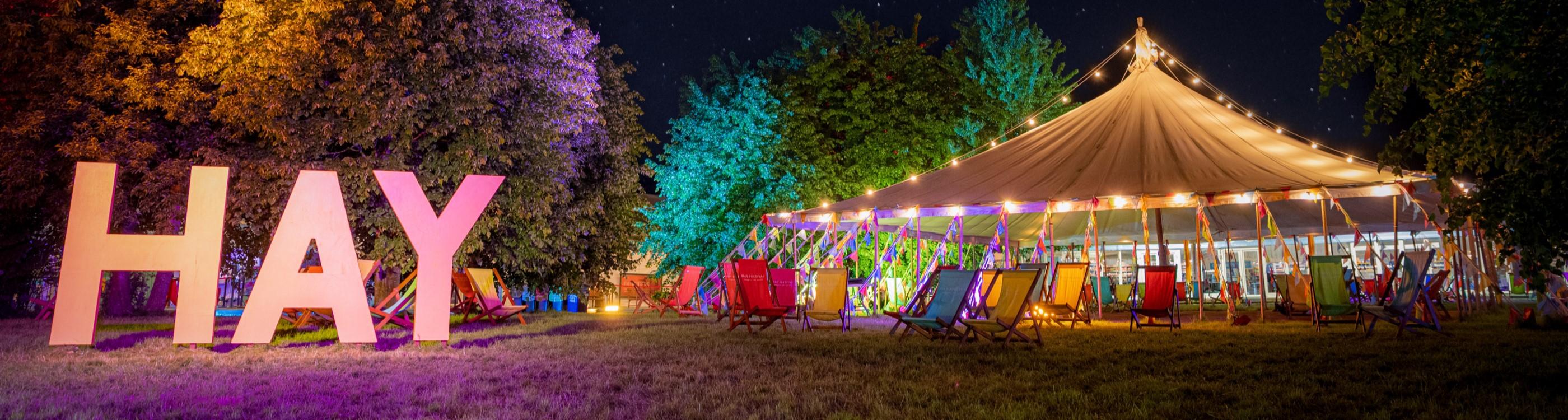 An illuminated tent with empty colourful deck chairs outside, at night, with a sign saying 'Hay', at Hay Festival.