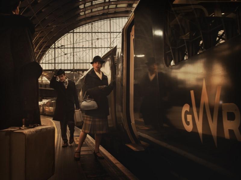 Stylised photo of two characters from Agatha Christie's The Mousetrap, dressed in period costume, boarding a GWR train.