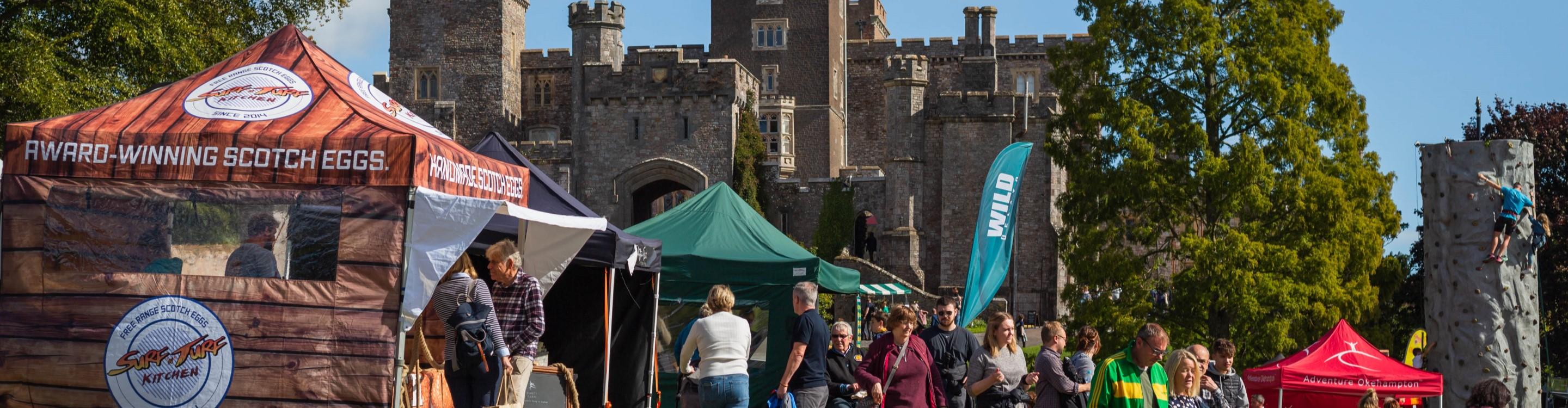 The Powderham Food Festival in full flow, with crowds of people in front of various food stalls and tents, with Powderham Castle in the background