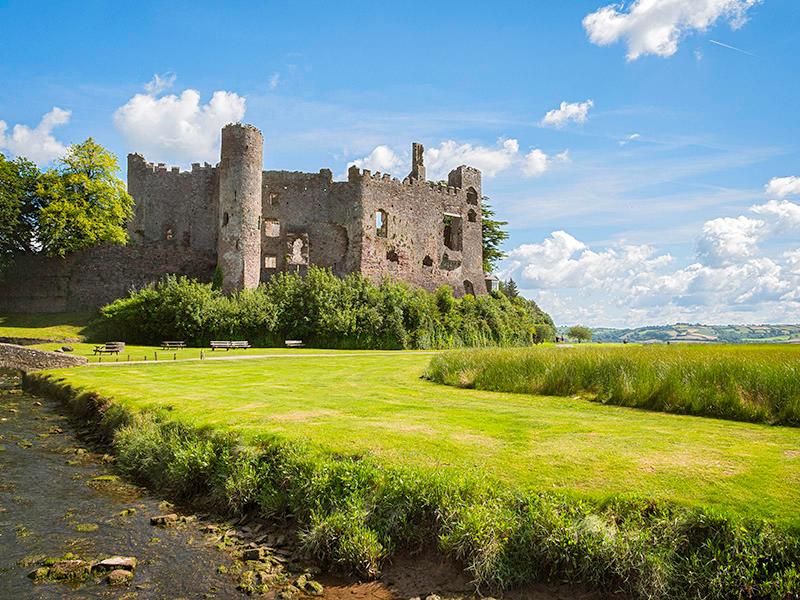 Long shot of Laugharne Castle on a brilliant day with clear blue sky in the background and a stream in the foreground