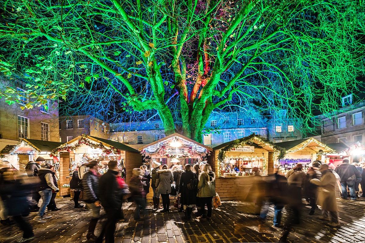 Stalls at Bath Christmas Market at Abbey Green, underneath a green illuminated tree