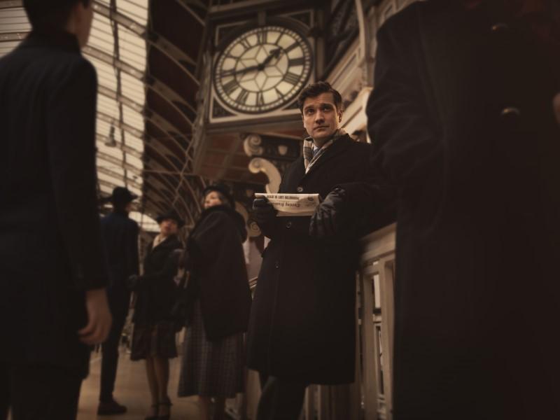 Stylised photo of a character from Agatha Christie's The Mousetrap, dressed in period costume, standing in front of a grand clock at a train station.